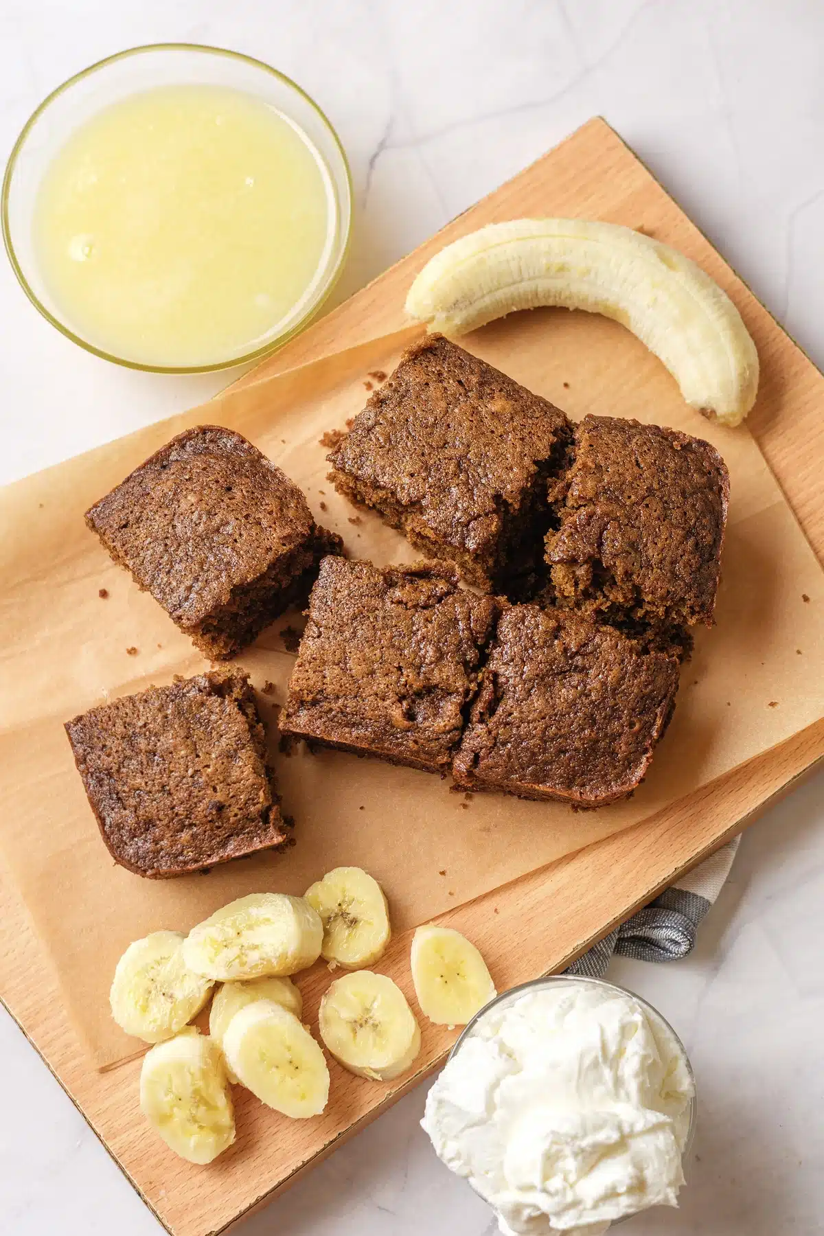 Gingerbread cake cut into squares on a parchment-lined board with sliced banana, whipped cream and a bowl of vanilla sauce