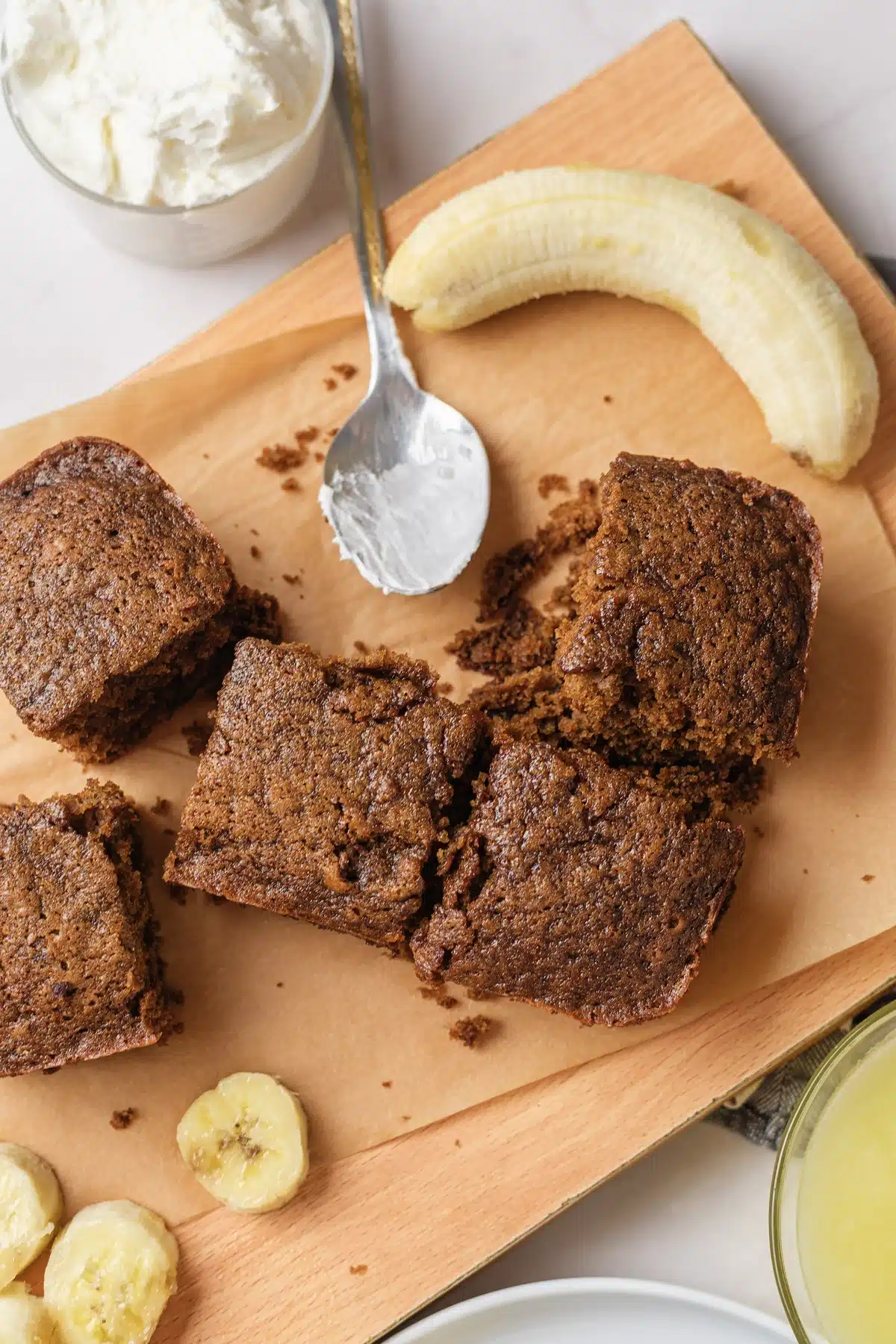 Dark spiced squares on a parchment-lined board with a tub of whipped cream, a banana and a spoon