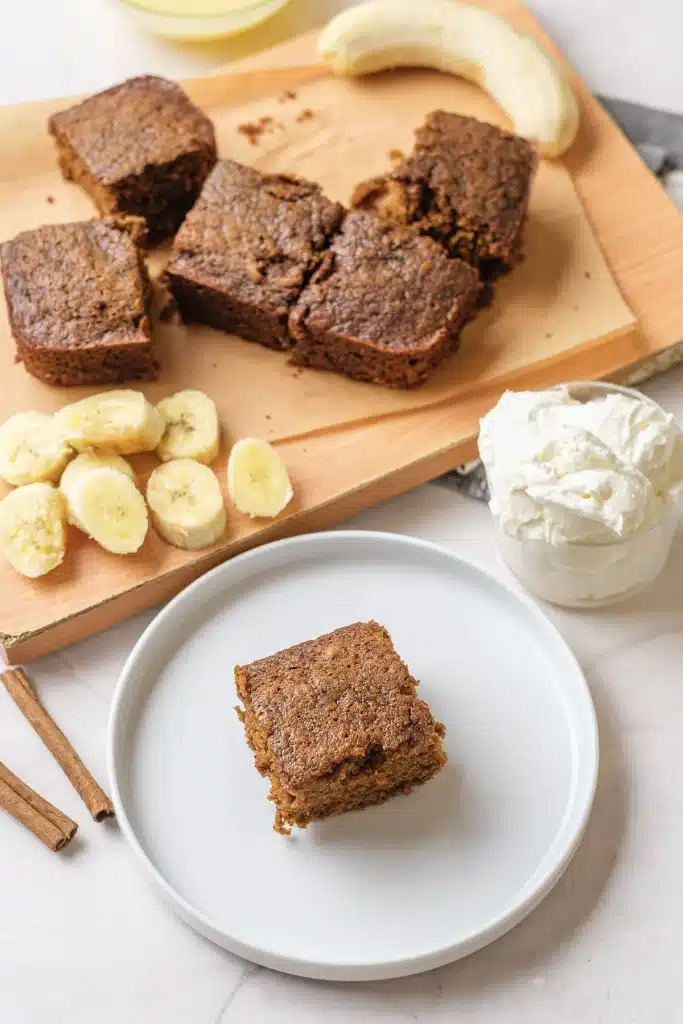 Gingerbread cake square on a white plate, seen from above, with the board of squares and sliced banana behind