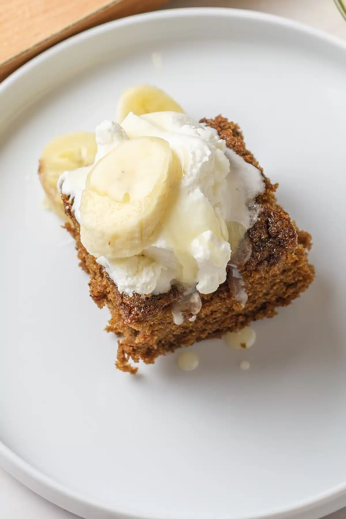 A close view of a dark spiced square topped with whipped cream and a slice of banana, with pale sauce running down the side