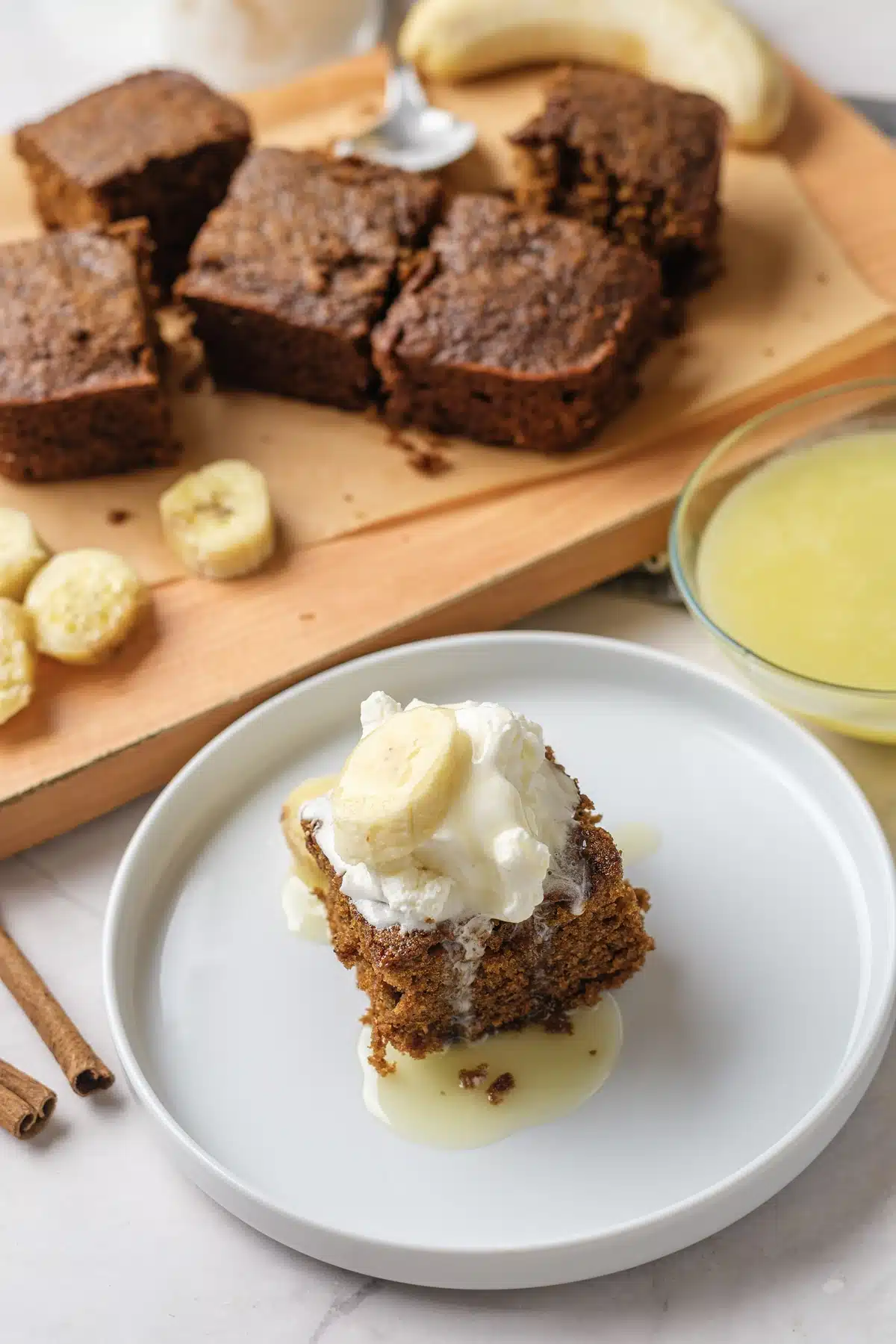 Gingerbread cake square on a white plate with whipped cream, banana and vanilla sauce, with the board of squares behind