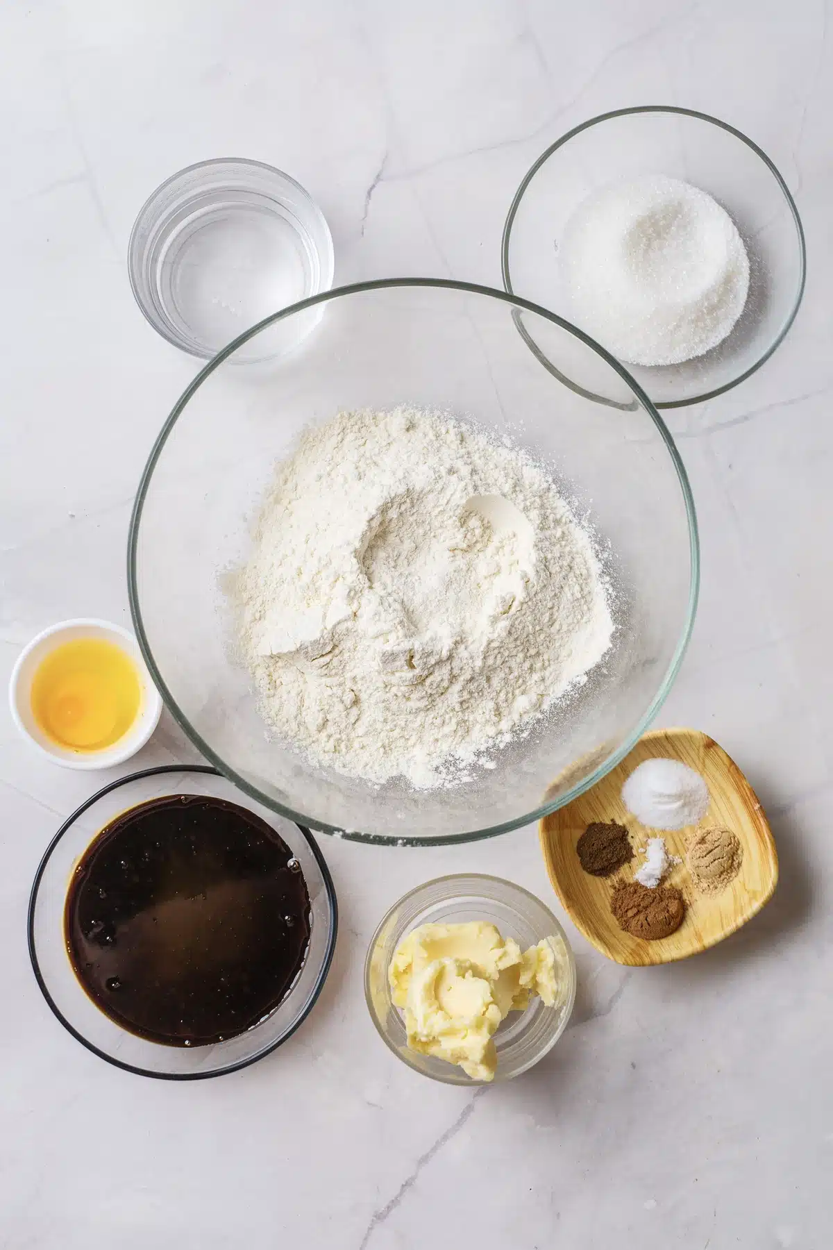 Ingredients for gingerbread cake: flour in a glass bowl, molasses, softened butter, an egg, granulated sugar, water and a dish of ground spices and leaveners