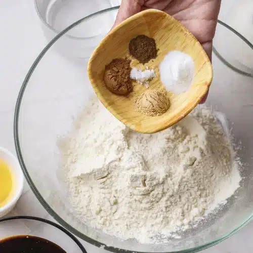 A wooden dish of ground spices and leaveners tipped onto flour in a glass bowl
