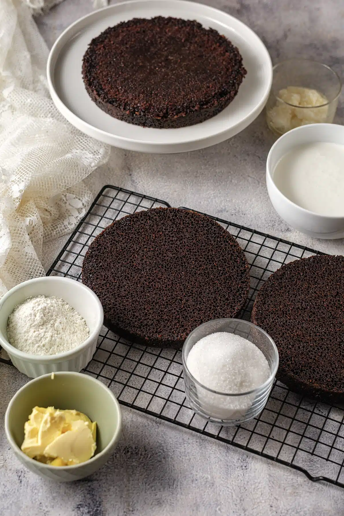 Two baked cocoa layers cooling on a rack beside bowls of flour, sugar, milk and butter