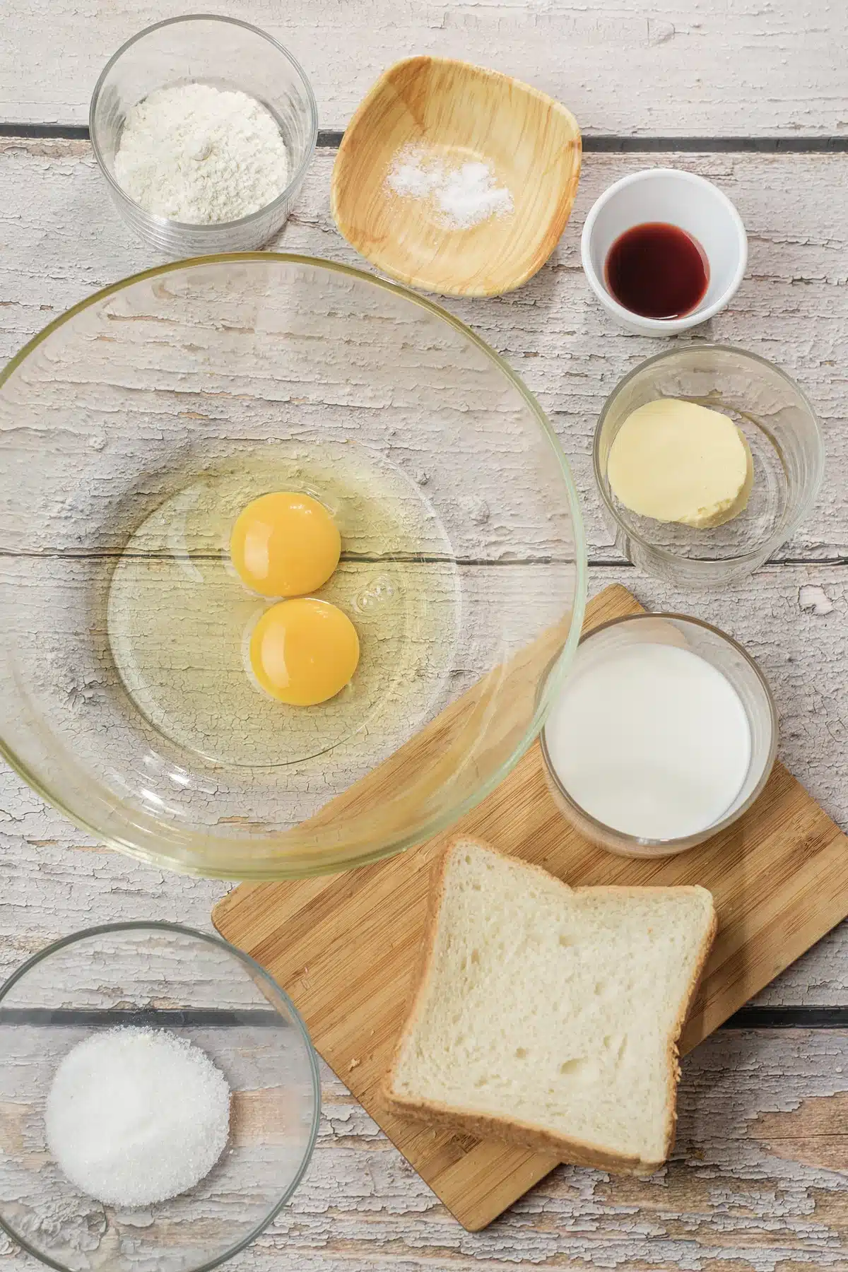 Eggs, milk, flour, sugar, vanilla and butter in small bowls beside thick white bread slices on a board