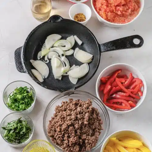 Sliced onion in a cast iron skillet with bowls of meat, peppers and herbs around it
