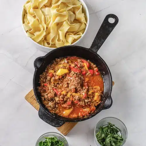The tomato and sausage sauce simmering in the skillet beside a bowl of cooked pasta