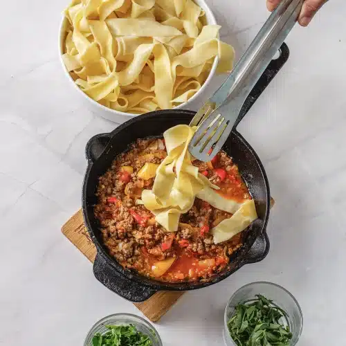 Tongs lifting wide ribbons of pasta into the simmering skillet