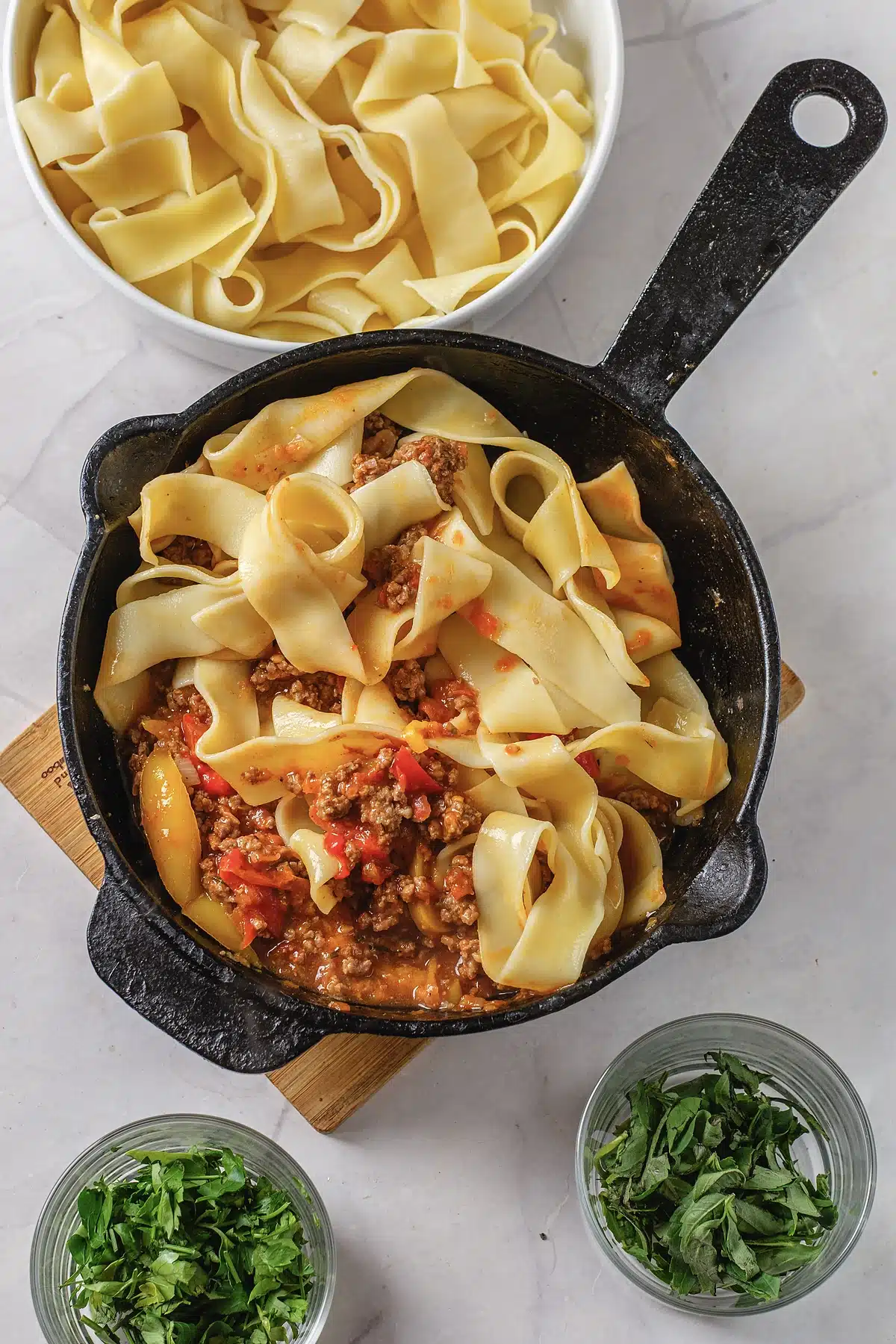 The finished skillet of ribbon pasta and sauce beside bowls of parsley and basil