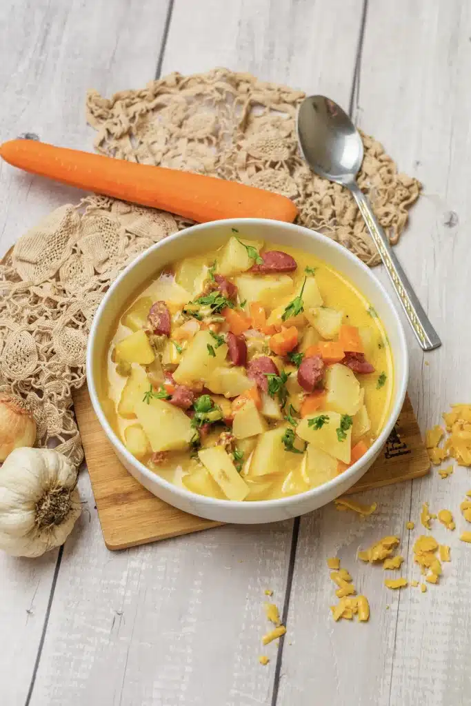 Kielbasa potato soup in a white bowl on a lace cloth with a whole carrot, a garlic bulb and grated cheddar around it