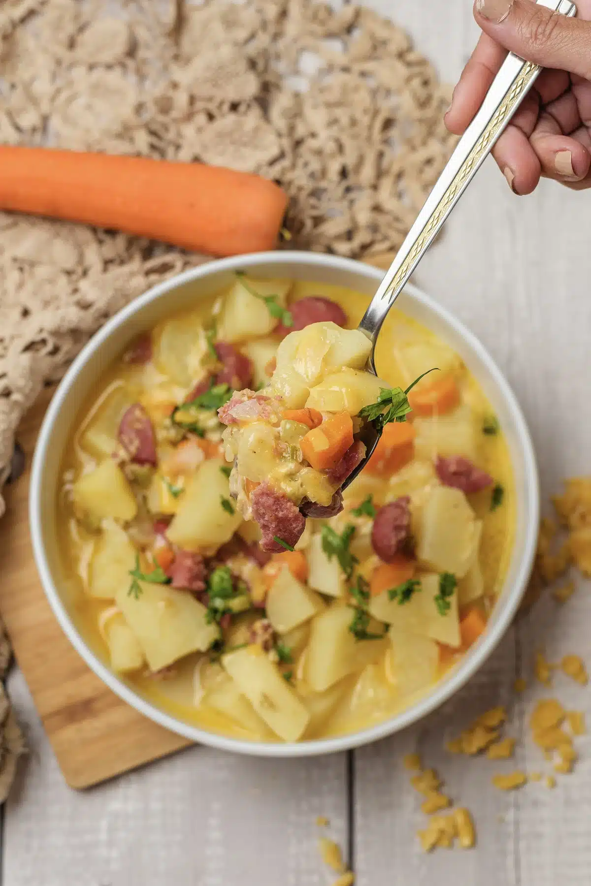 A hand lifting a spoonful of chunks and sausage pieces from a full white bowl on a lace cloth