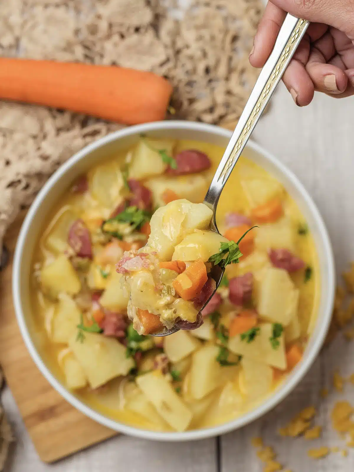 A spoon lifting sausage pieces, carrot and a chunk of Yukon Gold from a bowl, with grated cheese scattered on the table