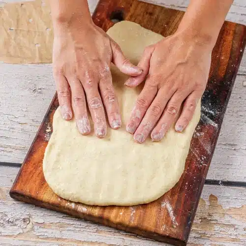 Two hands patting the soft dough flat on a floured wooden board