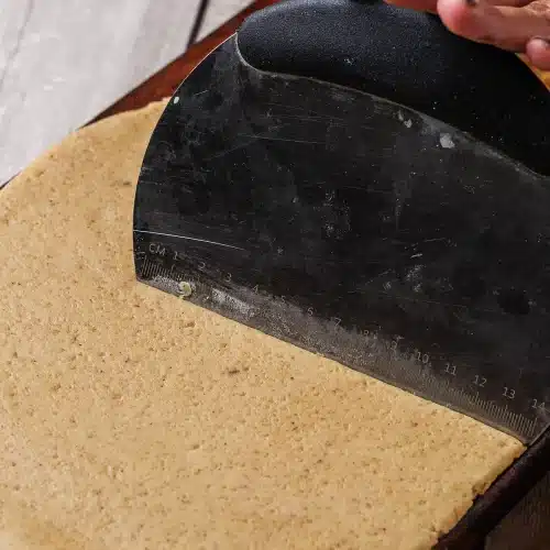A bench scraper lifting the edge of the rolled round on a wooden board