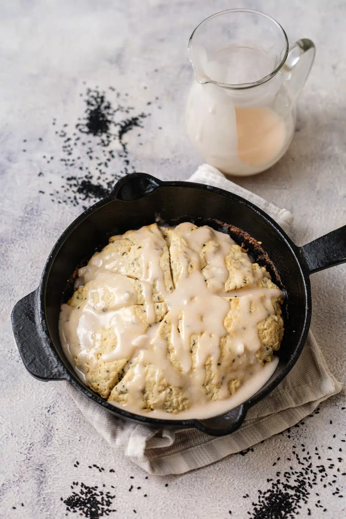 Lemon poppy seed scones baked as one round in a cast-iron skillet, glazed, with a jug of glaze behind