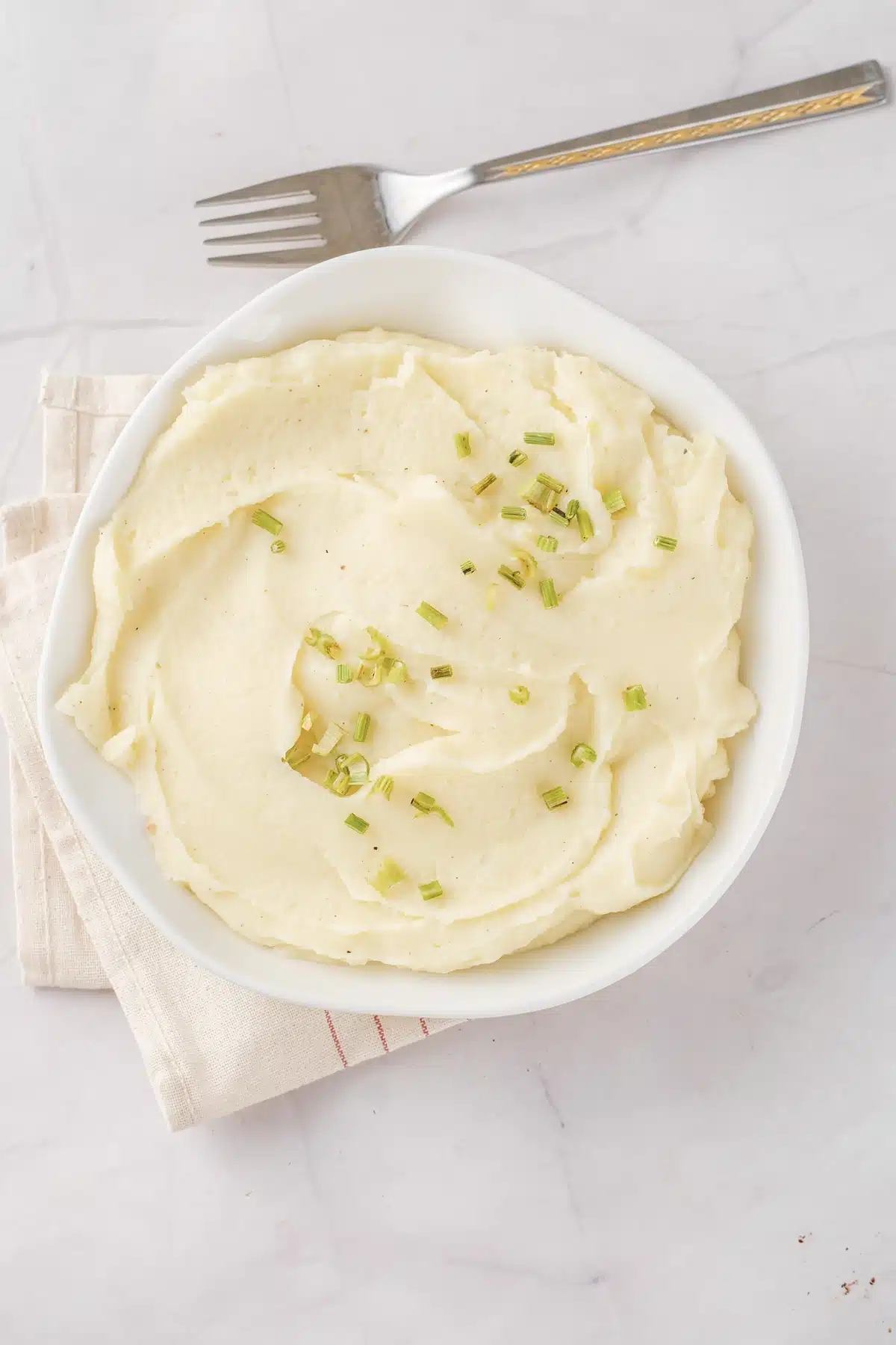 Mashed rutabaga in a white bowl with chives, seen from above with a fork and a striped cloth beneath