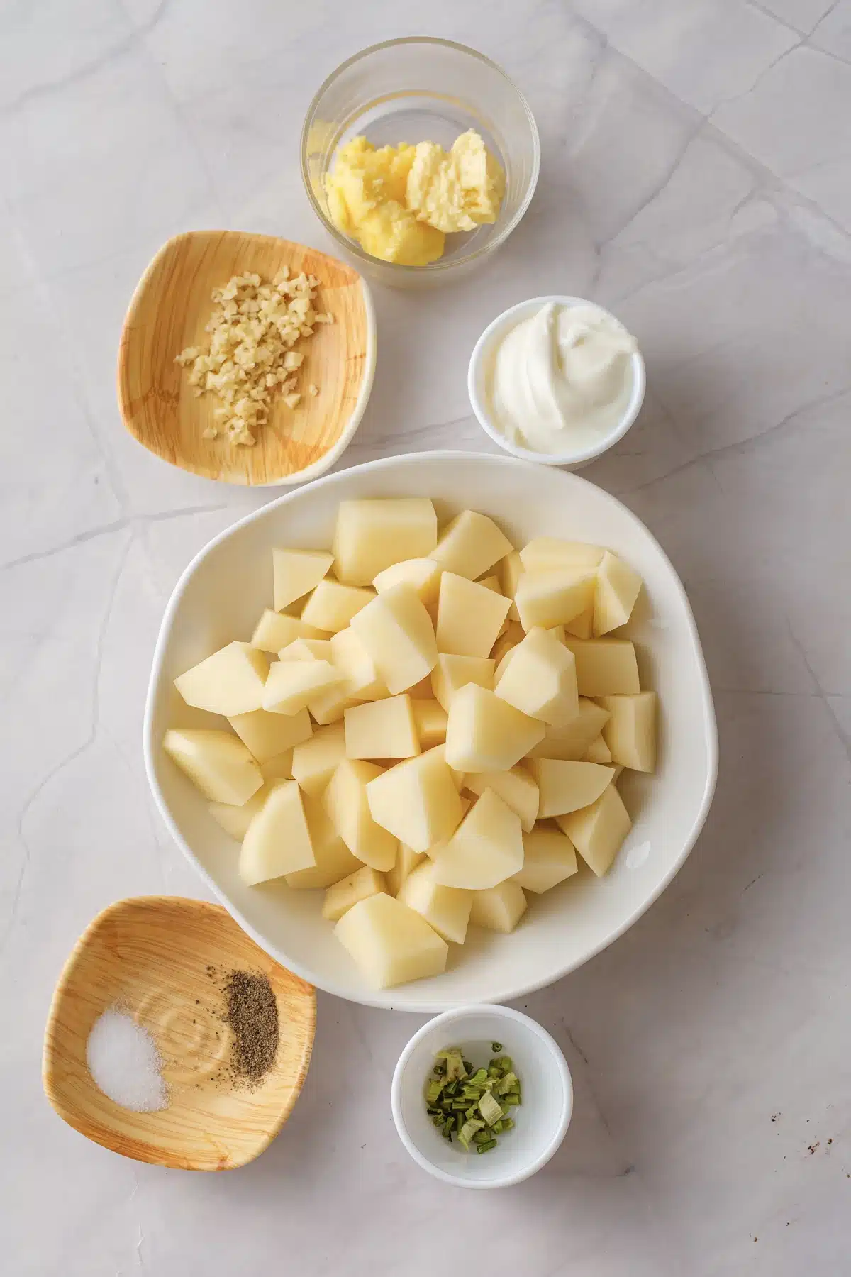 A bowl of peeled pale yellow cubes with butter, minced garlic, sour cream, salt, pepper and chopped chives around it