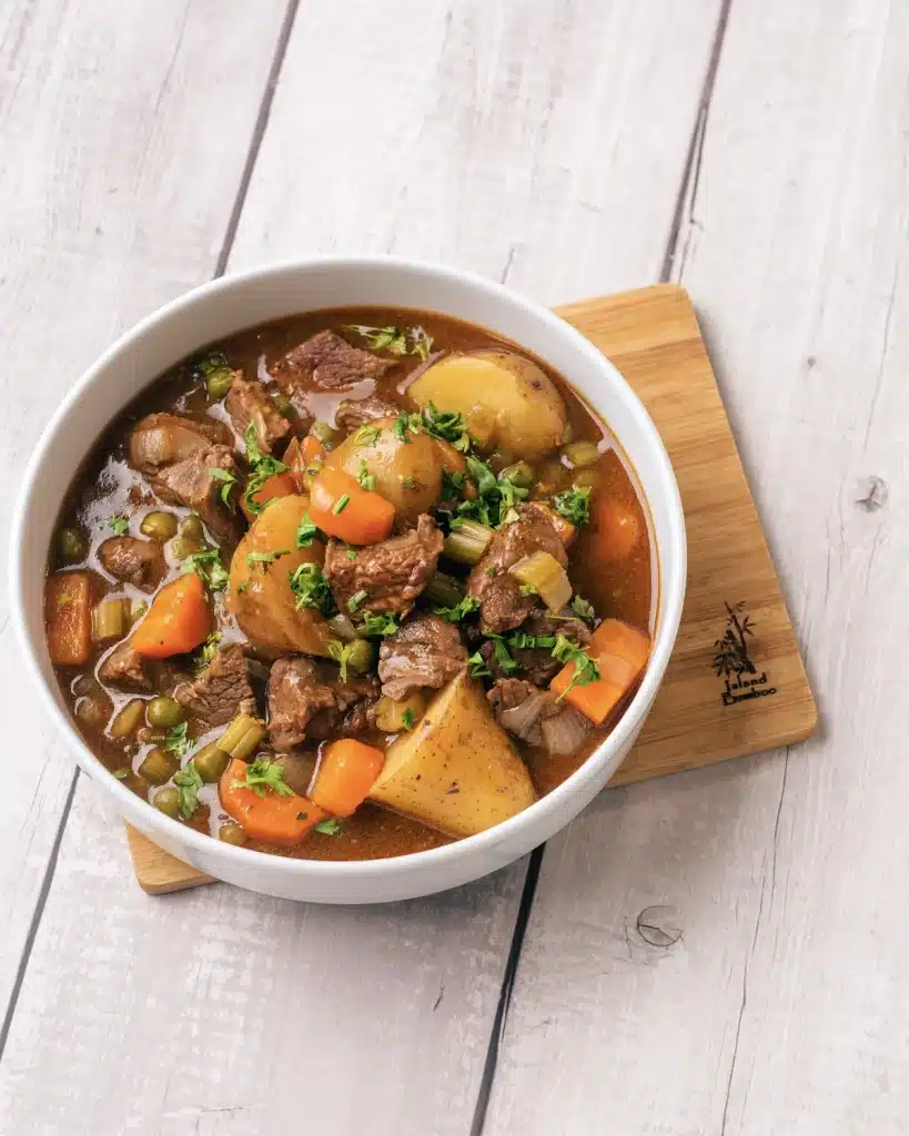 Homemade beef stew in a white bowl on a wooden board, seen from above, with potatoes, carrots, peas and parsley