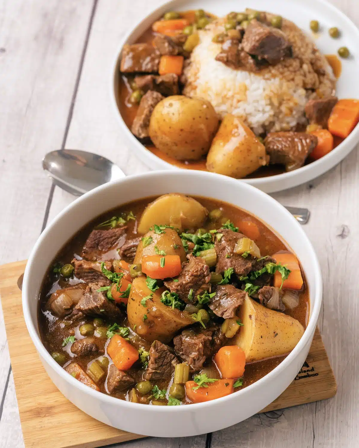 Homemade beef stew in a white bowl with big potato pieces, carrots and peas, and a plate of it over rice behind