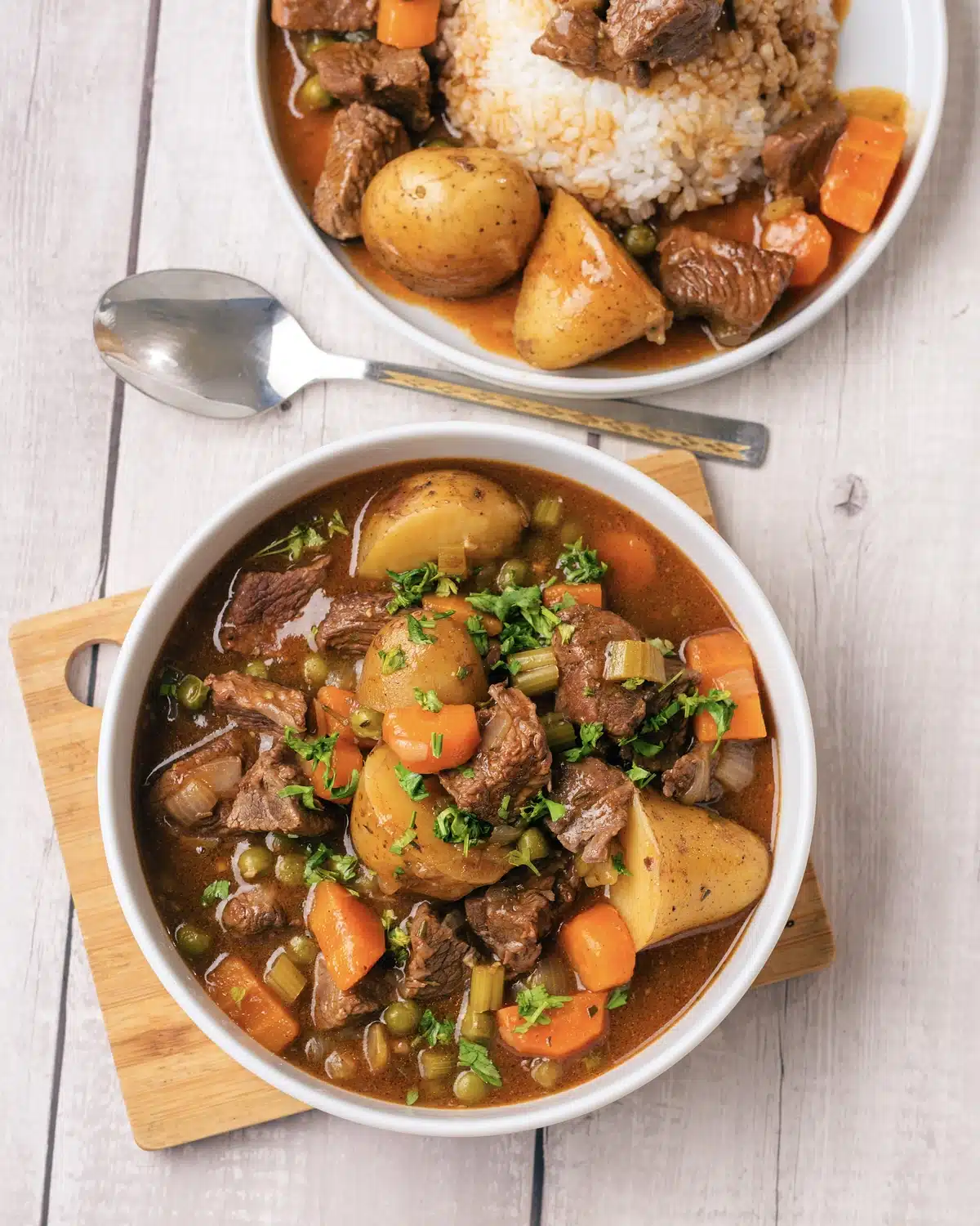 A bowl of chunky meat and vegetables in gravy on a board with a spoon and a plate of it over rice behind