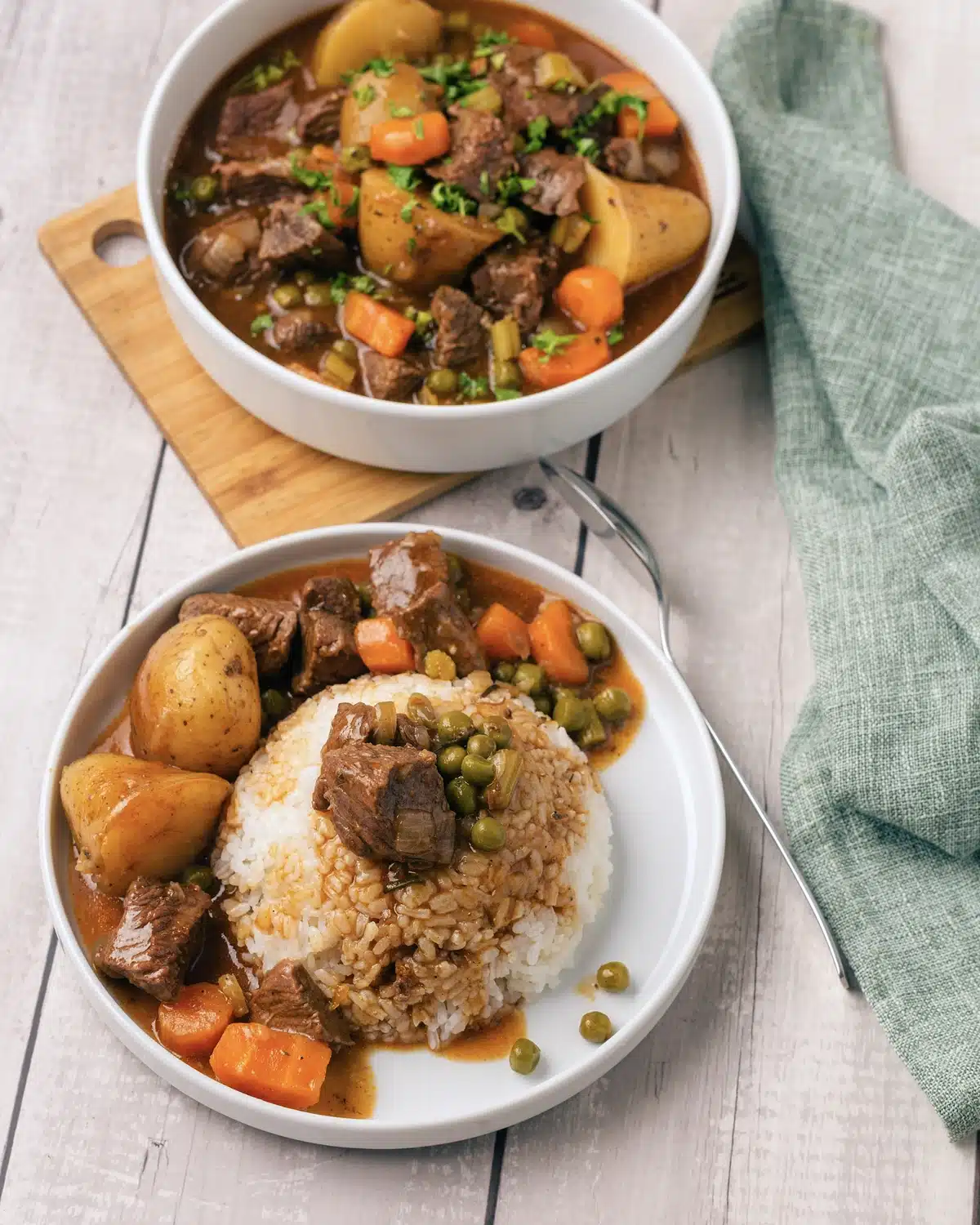 Homemade beef stew spooned over a mound of white rice on a plate, with a bowl of the stew and a green cloth behind