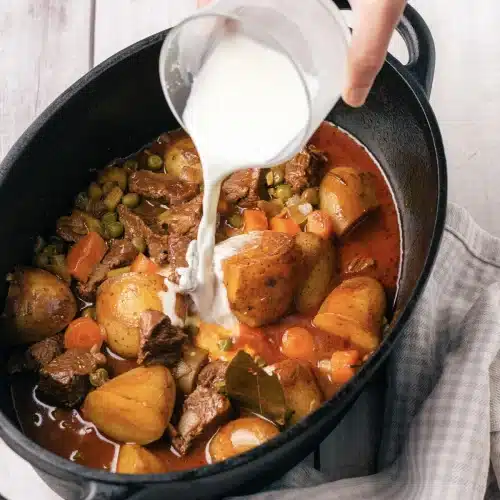 The cornstarch slurry poured from a cup into the pot of meat, potatoes, carrots and peas