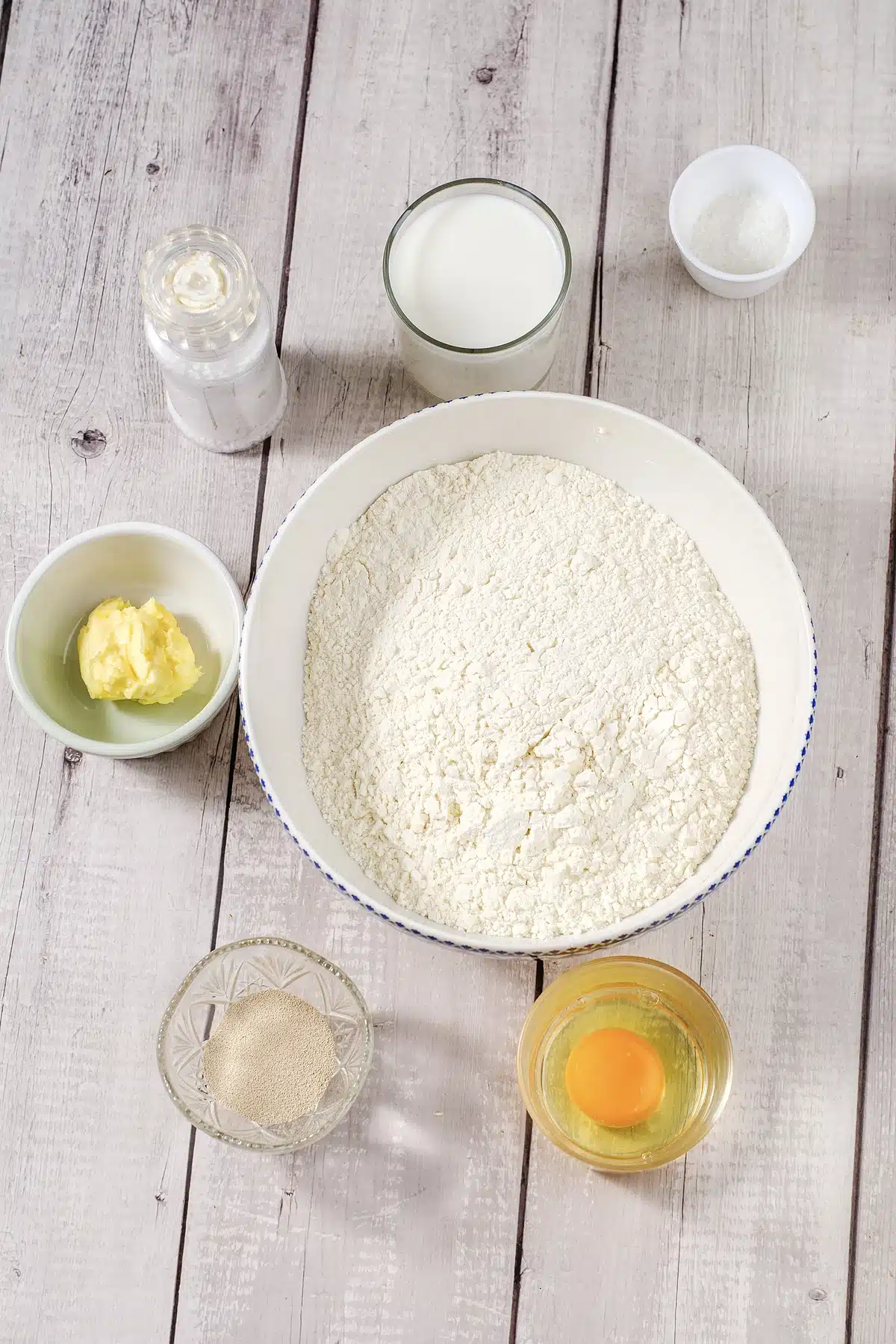 Flour, milk, melted butter, an egg, yeast, sugar and salt in bowls on white wood