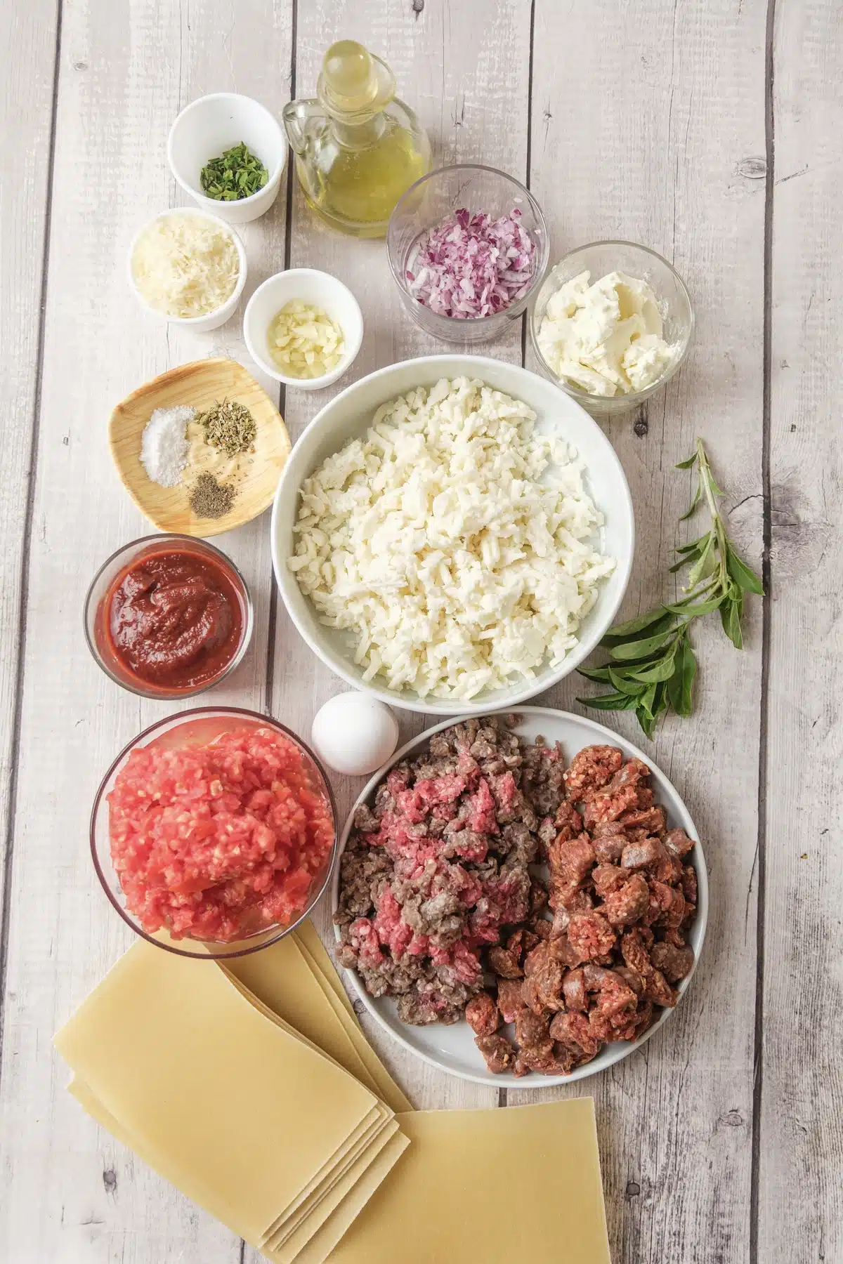 Ground beef, sausage, ricotta, shredded mozzarella, crushed tomatoes, red onion and flat noodles laid out on a board