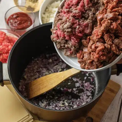 Raw ground beef and sausage tipped from a bowl into the pot of onion