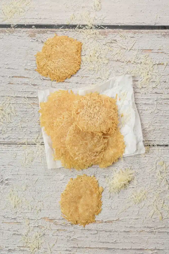 Parmesan crisps on a paper towel seen from directly above, golden and lacy, with loose shreds around them