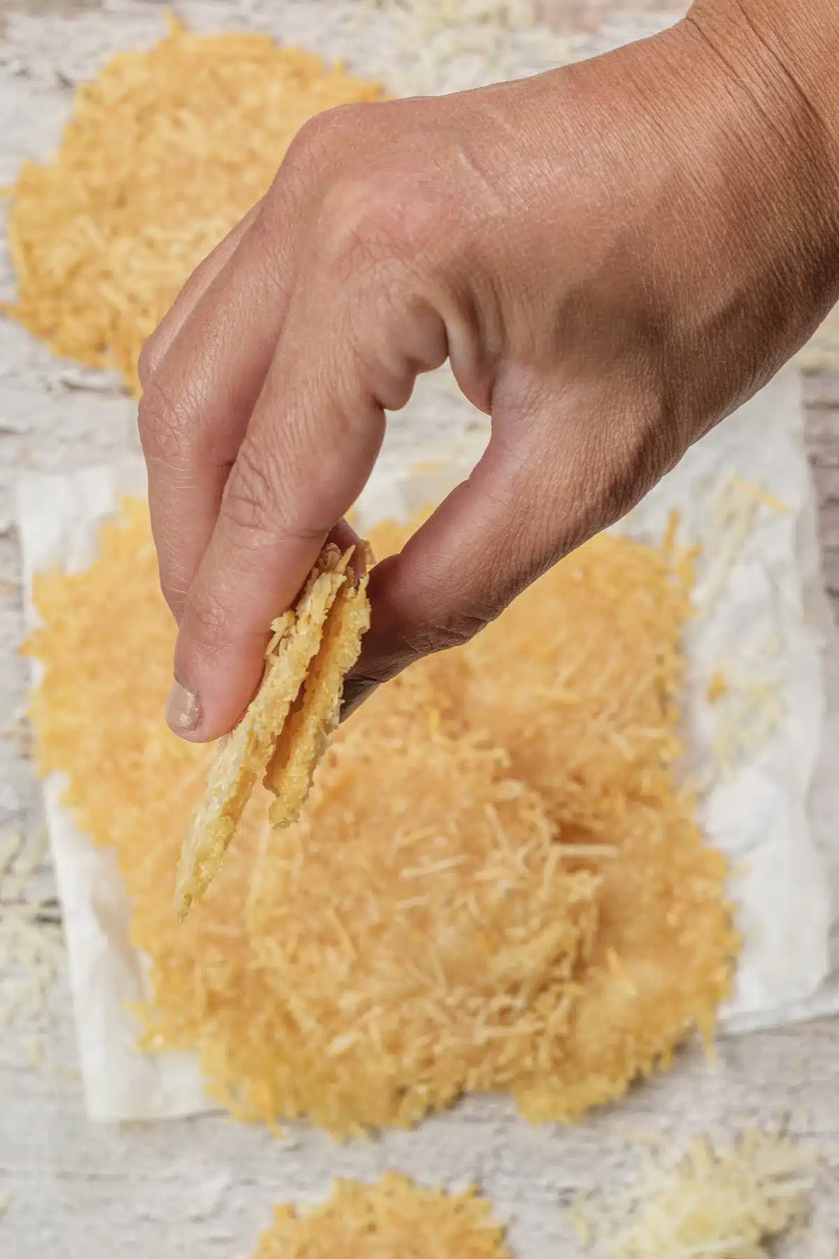 A hand lifting one Parmesan crisp off a stack on a paper towel, with shreds scattered around