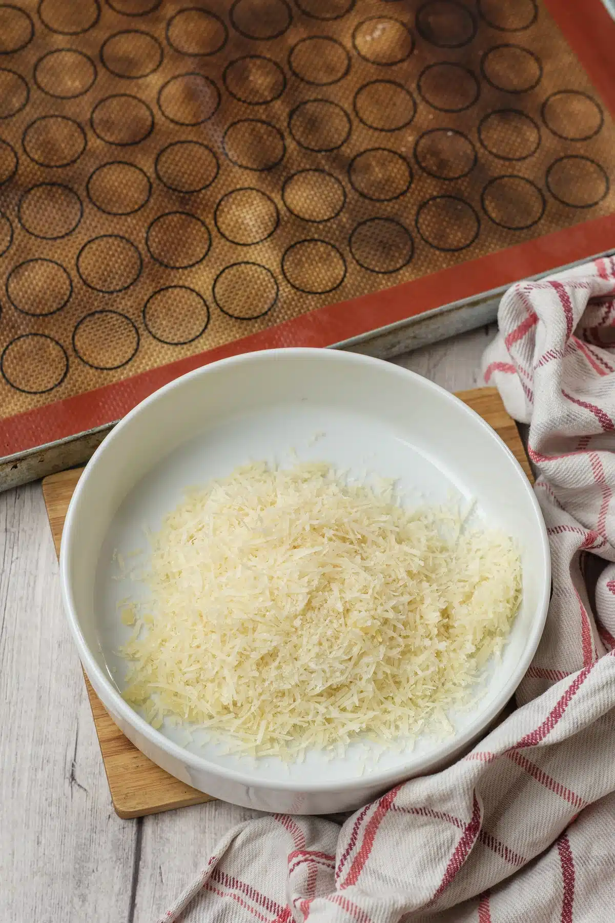 A white bowl of shredded hard cheese on a board beside a baking sheet lined with a silicone mat