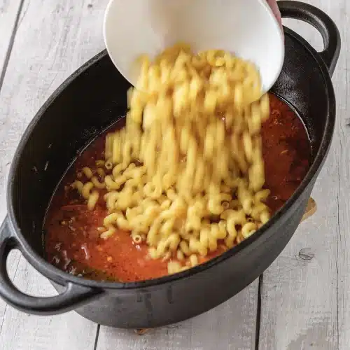Dry elbow macaroni poured from a bowl into the simmering sauce