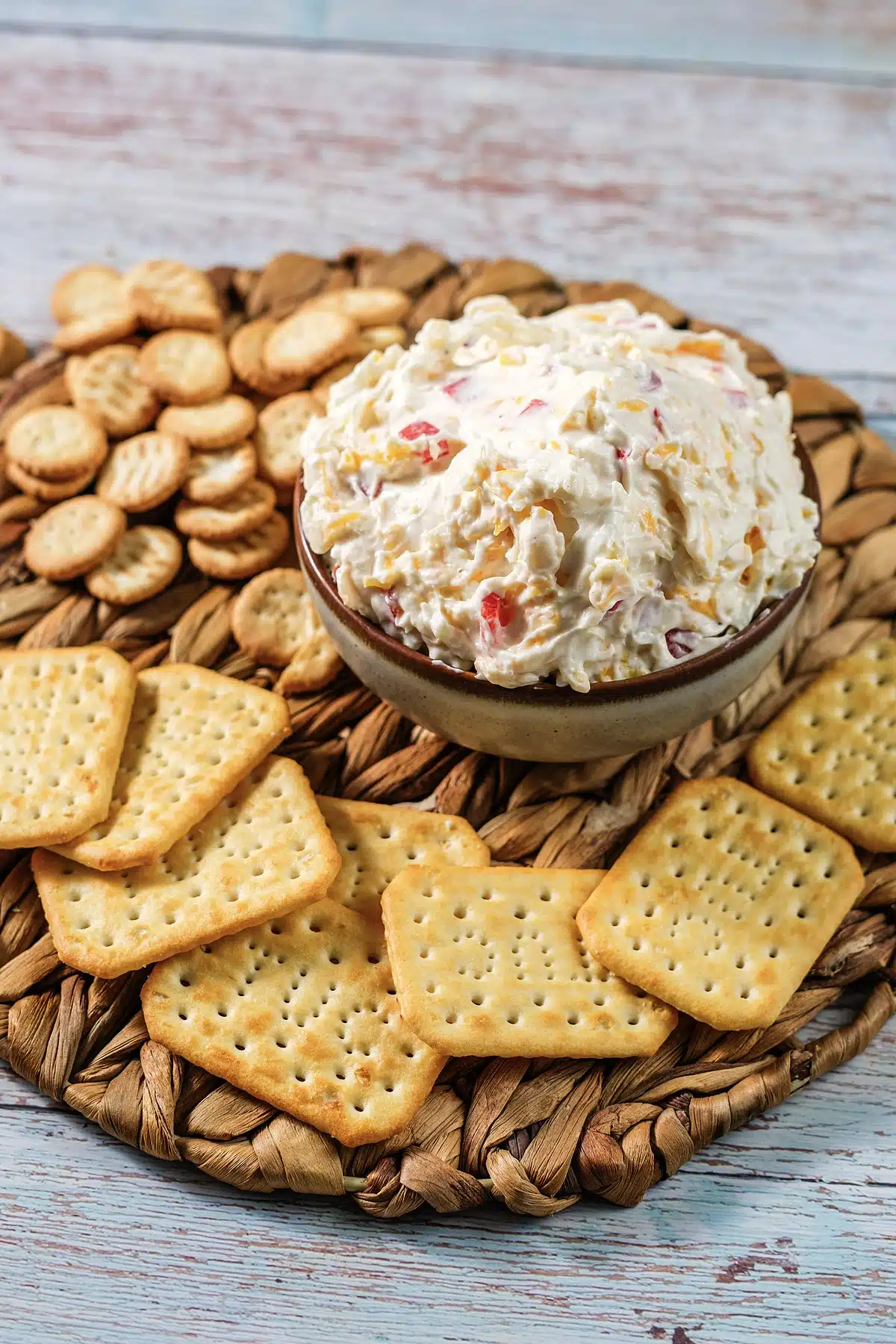 Pimento cheese in a small stoneware bowl with crackers scattered on a woven mat