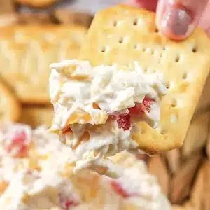 One square cracker loaded with the creamy spread and held above the bowl