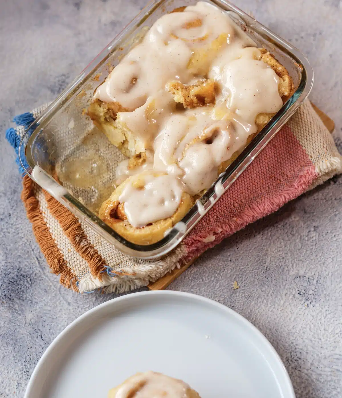 Pumpkin cinnamon rolls frosted in a rectangular glass dish on a striped cloth with a white plate beside it