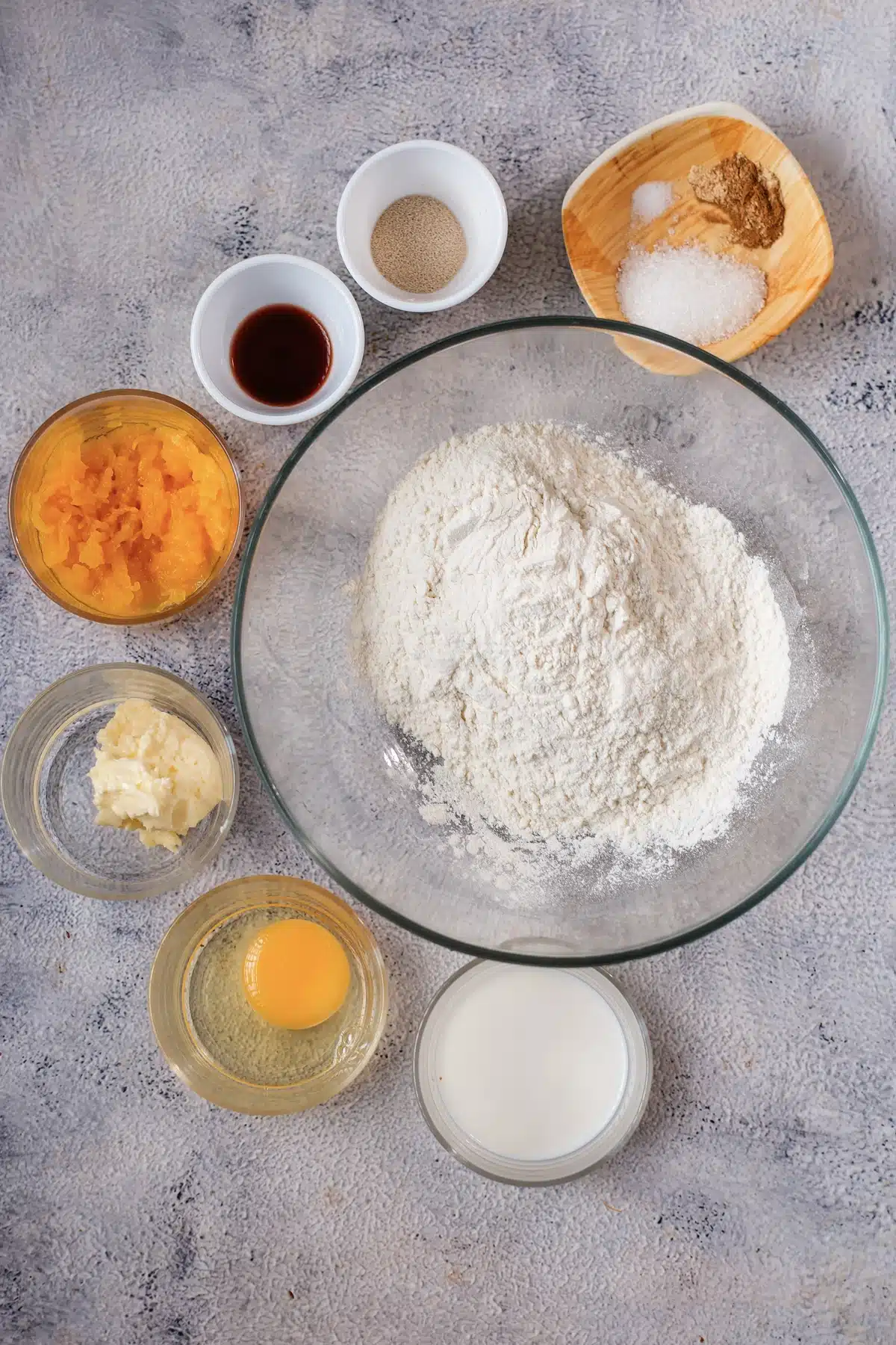 Flour, pumpkin puree, an egg, milk, melted butter, vanilla, yeast, salt and spice laid out on a gray surface