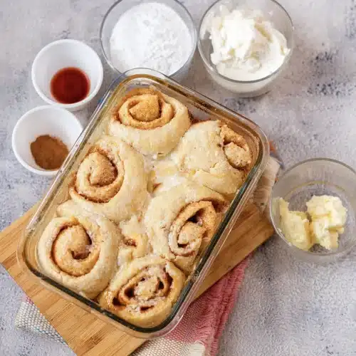 Risen, unbaked rolls in a glass dish with cream cheese, powdered sugar, butter and vanilla arranged around it