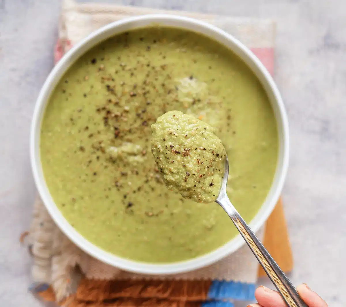 A spoonful of smooth green puree lifted over a white bowl on a striped napkin