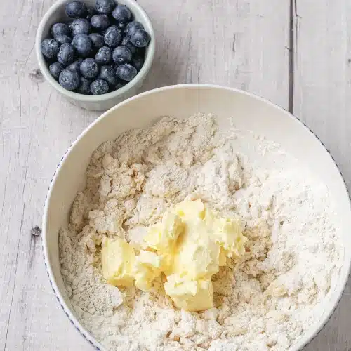 Cubes of cold butter dropped onto flour in a white bowl beside fresh fruit