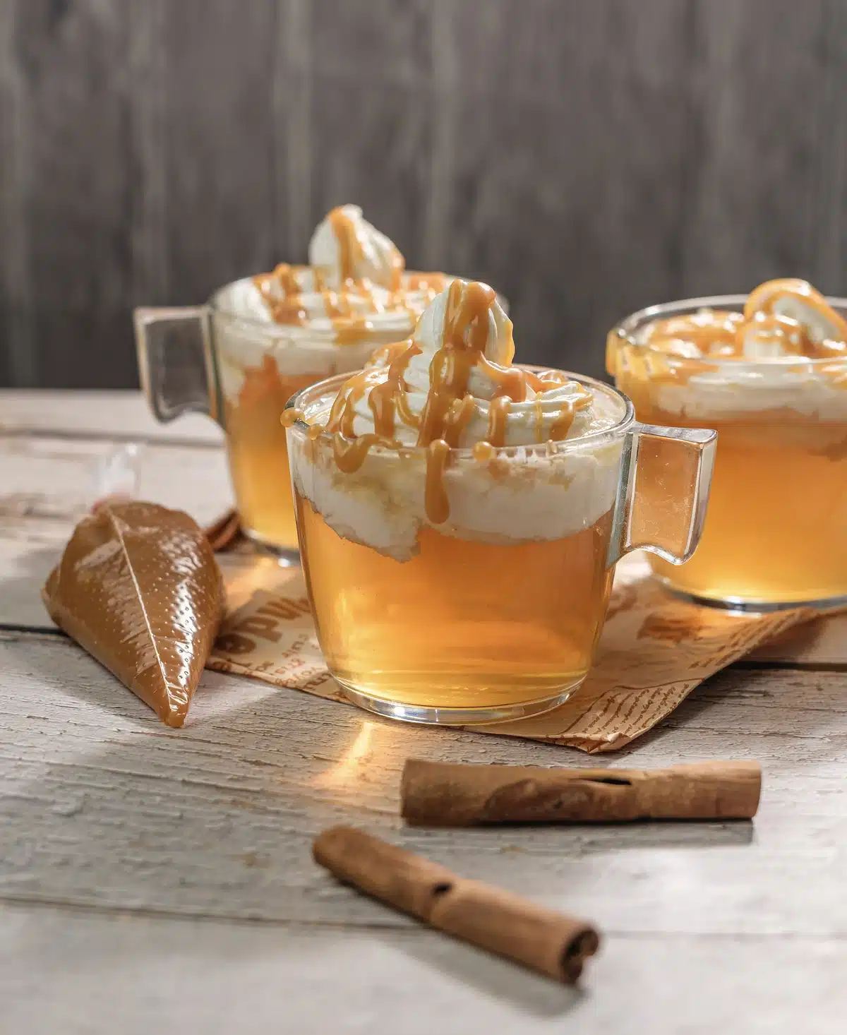 Three glass mugs of Starbucks apple cider with whipped cream and caramel drizzle beside a piping bag of caramel