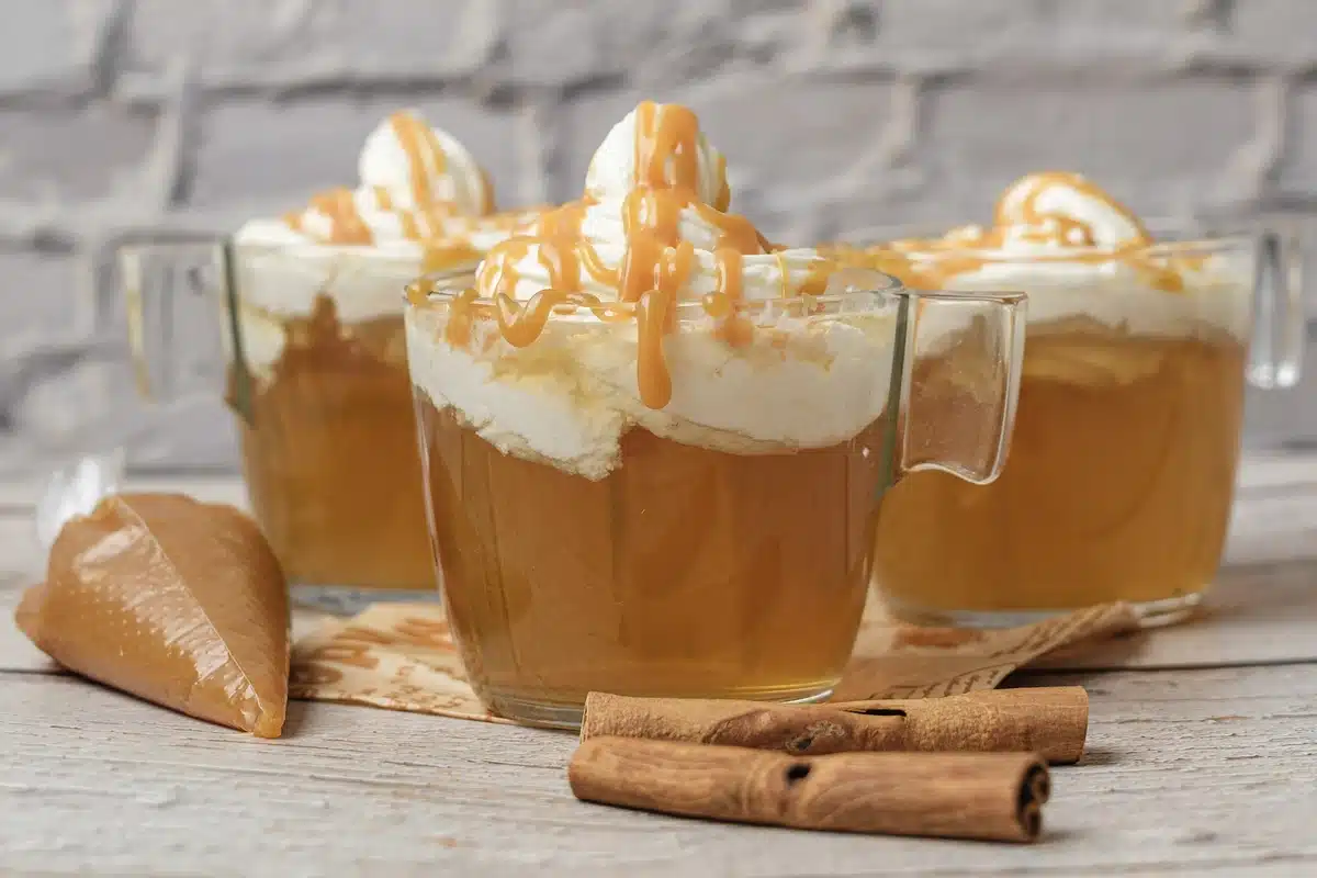 Three glass mugs of Starbucks apple cider in a row against a white brick wall with cinnamon sticks in front