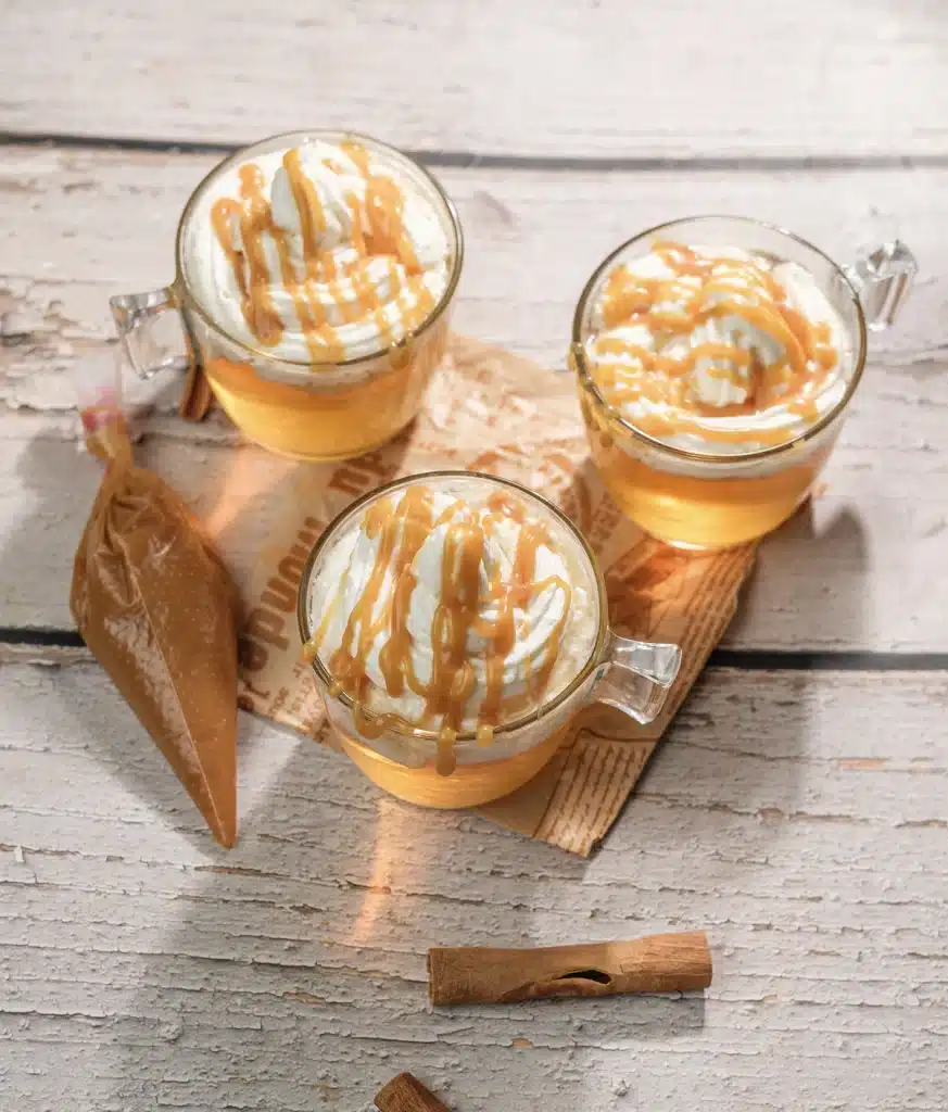 Starbucks apple cider in three glass mugs seen from above, topped with whipped cream and caramel drizzle