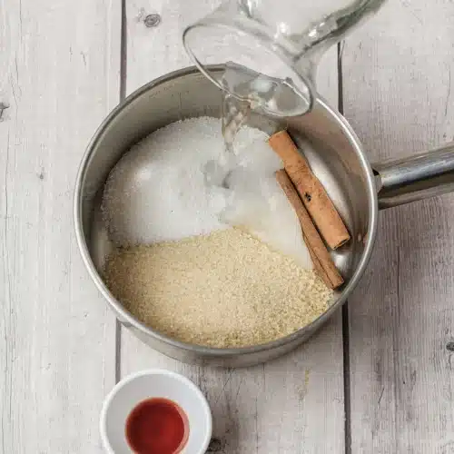 Water poured from a pitcher over the sugars and cinnamon stick in the saucepan