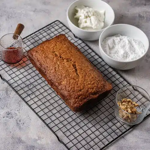 The baked cake cooling on a wire rack beside bowls of cream cheese and powdered sugar