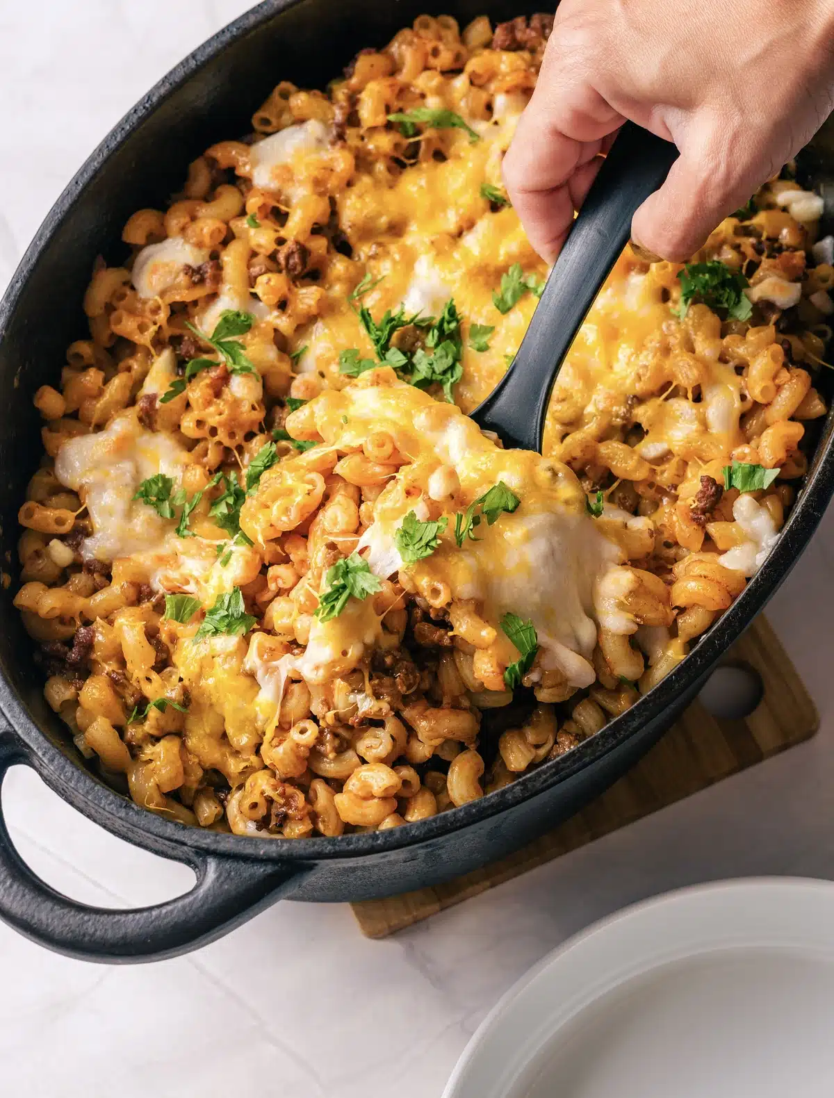 Homemade beefaroni lifted on a black spoon from a cast-iron pot, with melted cheese stretching and chopped parsley on top