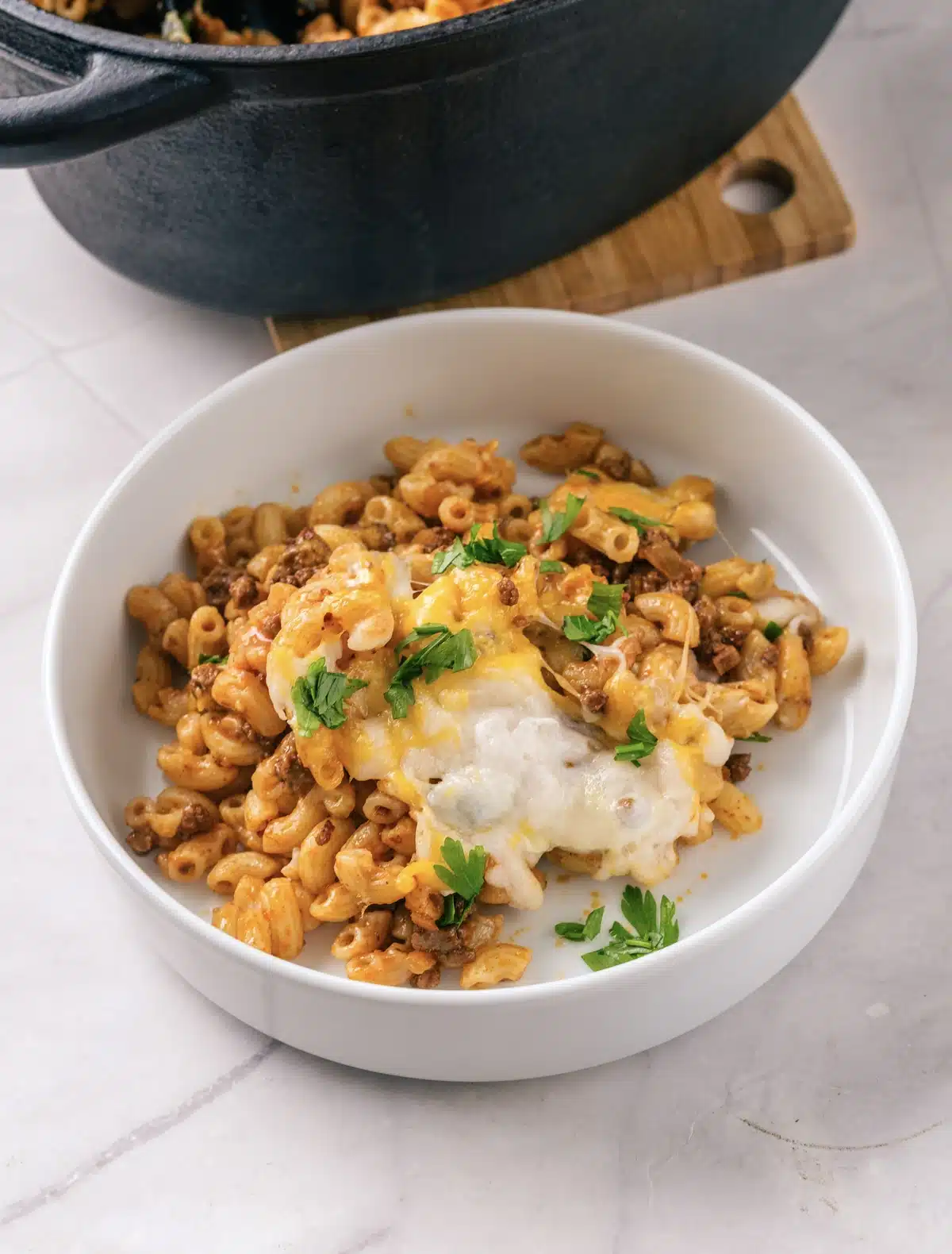 A bowl of homemade beefaroni with melted cheese and parsley on a marble counter, with the cast-iron pot behind
