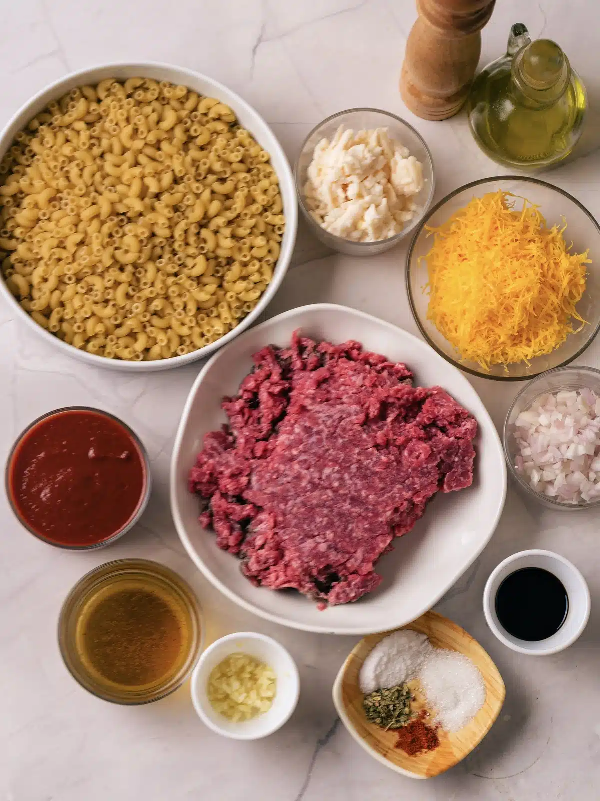 Dry corkscrew pasta, ground beef, tomato sauce, shredded cheddar, mozzarella, onion, garlic, broth, oil and spices laid out on a marble surface