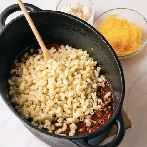 The pasta stirred into the sauce with a wooden spoon, with the cheeses waiting beside the pot
