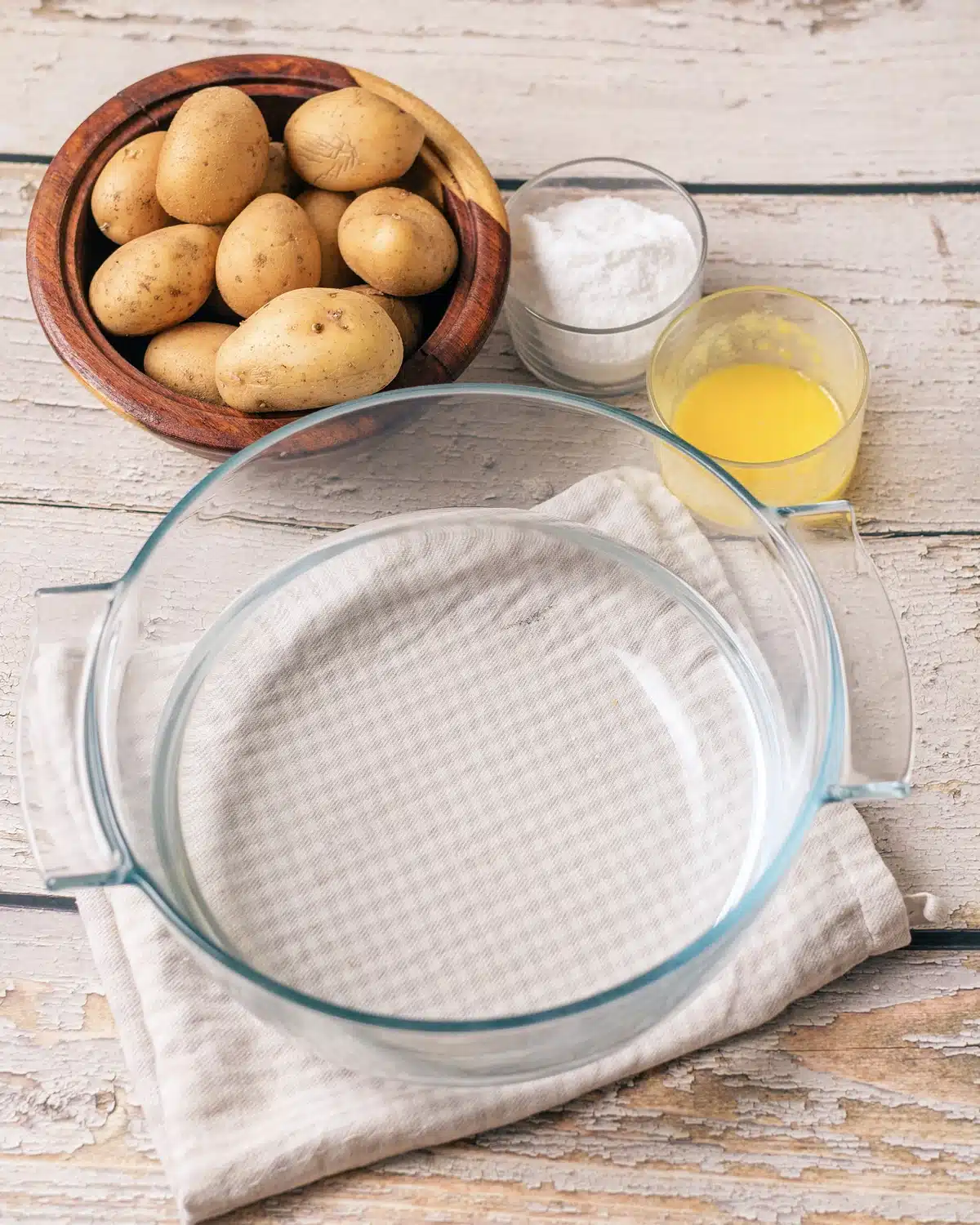 Small yellow spuds in a wooden bowl beside a dish of fine white seasoning, a glass of melted butter and an empty glass dish